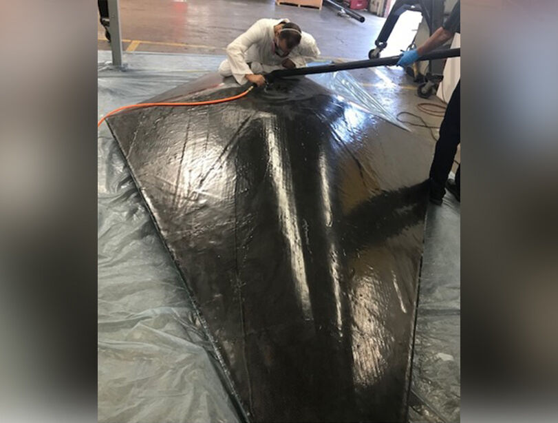 Images of warehouse space with the test body laying on a protective surface on the floor. Technicians are using 
a tool on the carbon fiber surface of RF test body.
